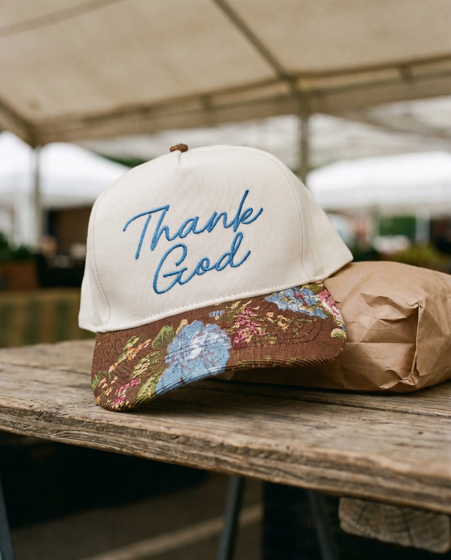 Thank God Script Hat with floral tapestry brim resting on a weathered farmers market table